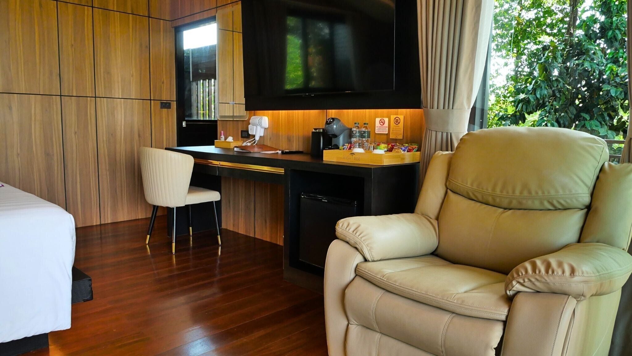 A hotel room with a bed, a desk with a chair, a leather armchair, a television, and a small refrigerator.