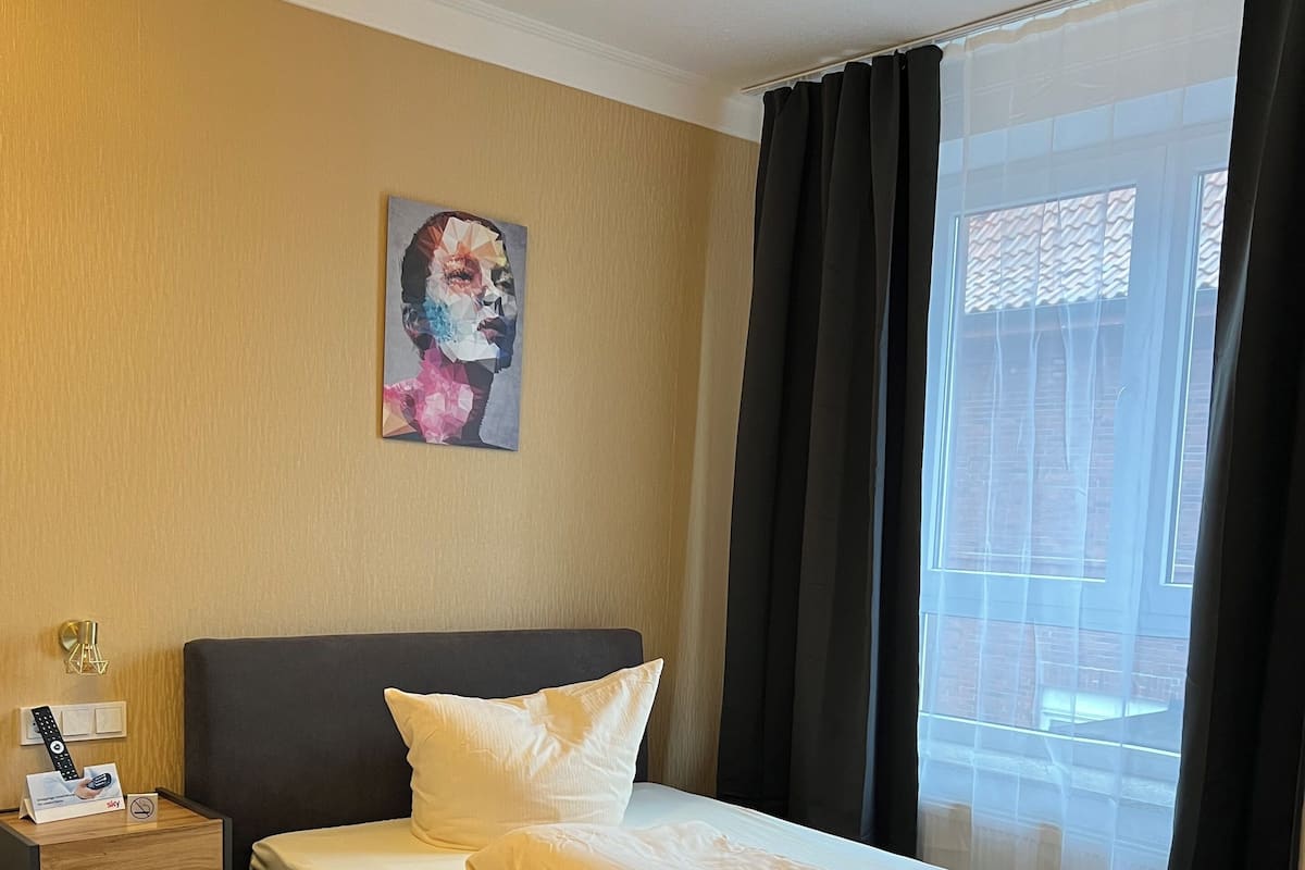 A hotel room with a bed, a nightstand with a remote control, a painting on the wall, and a window with curtains.