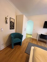 Una habitación compacta con un sillón verde azulado, un armario blanco, un escritorio y un televisor.