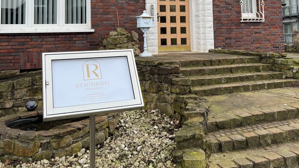 A brick building with a wooden door and multiple windows, a stone staircase, and a signboard. - Stadthotel Reckers (Werne)
