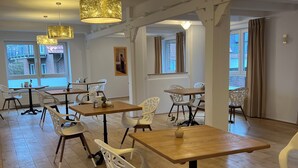 A spacious dining area with wooden tables and white chairs, large gold-toned pendant lights, and large windows with curtains. - Stadthotel Reckers (Werne)