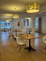 Un comedor amplio con mesas de madera y sillas blancas, grandes lámparas colgantes doradas y ventanas grandes con cortinas.