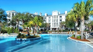 Una gran piscina curva con tumbonas y palmeras, rodeada por un hotel de varios pisos con balcones y un cielo azul despejado.