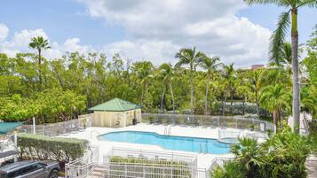 Una piscina con una valla blanca, rodeada de palmeras y un pequeño edificio con techo verde.