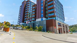 A modern multi-story building with a mix of glass and brown facade, featuring balconies and a parking area in front. - Catalina Bay Seascape with Bay Views and Carpark (Auckland West)