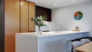 A modern kitchen with a central island, wooden cabinets, a white countertop, and a wall-mounted clock. - Catalina Bay Seascape with Bay Views and Carpark (Auckland West)