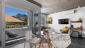 A modern living room with a glass door leading to a balcony, a round table with chairs, a sofa, a TV, and a small desk with a plant. - Seaside Studio Perfect Location (Cape Town)