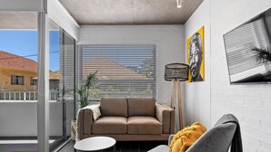 A modern living room with a sofa, armchair, and a small table. There is a large window with blinds, a potted plant, and artwork on the walls. - Seaside Studio Perfect Location (Cape Town)