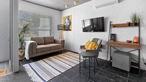 A modern living room with a sofa, a chair, a coffee table, a TV, and a desk with a plant and a salt lamp. - Seaside Studio Perfect Location (Cape Town)