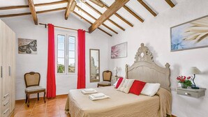 A bedroom with a wooden ceiling, a bed with a beige bedspread, a nightstand with a lamp, and a chair. - Villa Verde Ciutadella by Villa Plus (Ciutadella de Menorca)