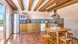 A kitchen with wooden cabinets, a built-in oven, a microwave, and a coffee maker. There is a dining table with chairs and a window overlooking a garden. - Villa Verde Ciutadella by Villa Plus (Ciutadella de Menorca)