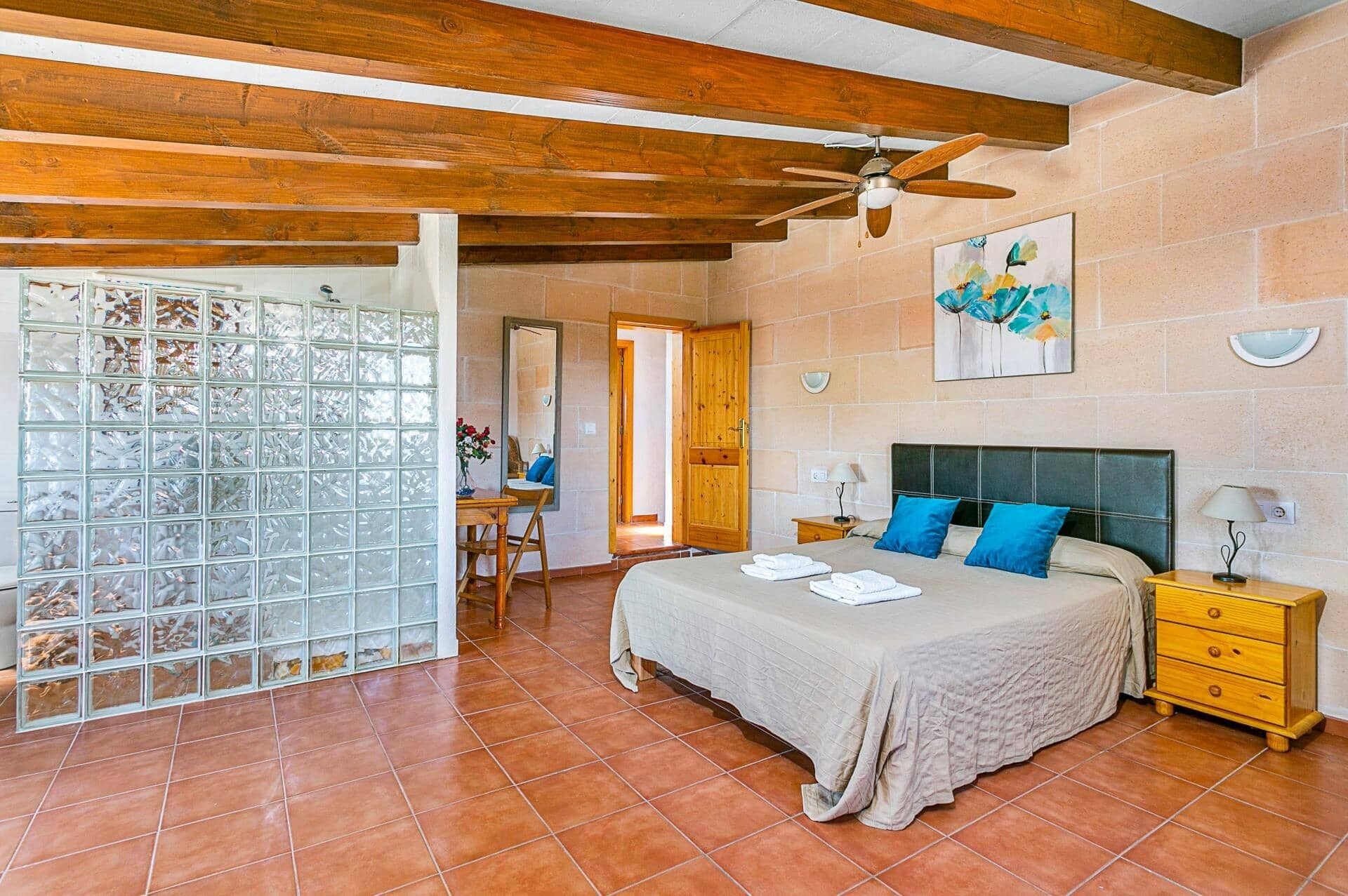 A bedroom with a bed, nightstands, a wooden ceiling fan, a bathroom with a glass door, and a tiled floor.