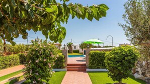 A pathway leading to a pool area with a stone wall, a palm tree, and a green umbrella. - Villa Verde Ciutadella by Villa Plus (Ciutadella de Menorca)