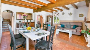 A spacious living area with a dining table set for four, a sofa, and a coffee table. There are wooden beams on the ceiling, a staircase, and a view of the outdoors through a window. - Villa Verde Ciutadella by Villa Plus (Ciutadella de Menorca)
