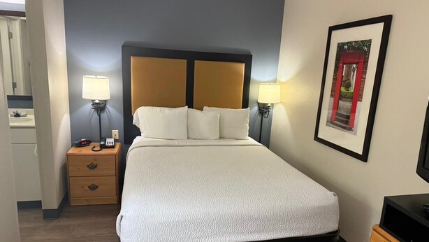 A hotel room with a bed, bedside lamps, a nightstand, a framed picture on the wall, and a television on a stand.