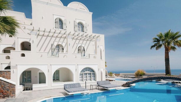A pool area with a clear view of the ocean, a white building with arches, and a palm tree. - Blue Arise Fira (Santorini)