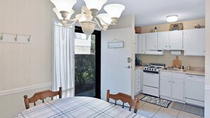 A room with a dining table, chairs, a chandelier, a kitchen with white cabinets, and a view through a door.