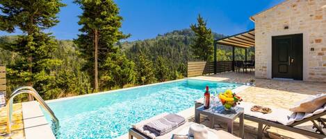 A poolside area with a stone building, lounge chairs, and a table with a bottle and fruit.