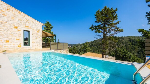 Una piscina junto a un edificio de piedra con una pérgola, rodeada de árboles y colinas.