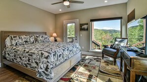 Un dormitorio con cama, escritorio, silla, ventilador de techo, ventana con vista a árboles y una puerta.