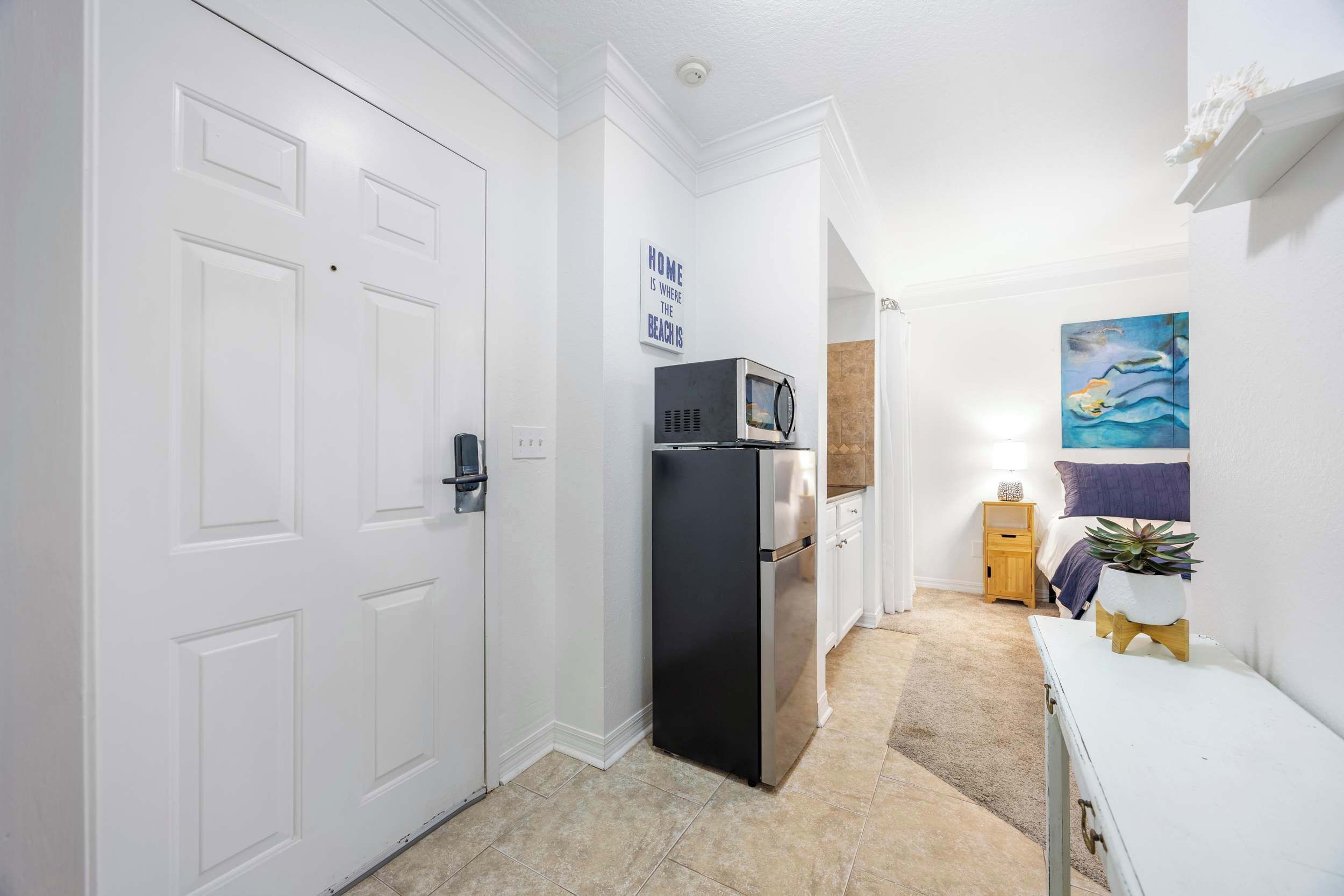A compact kitchen with white cabinetry, a black refrigerator, and a microwave. A small wooden nightstand with a lamp and a potted plant is visible.