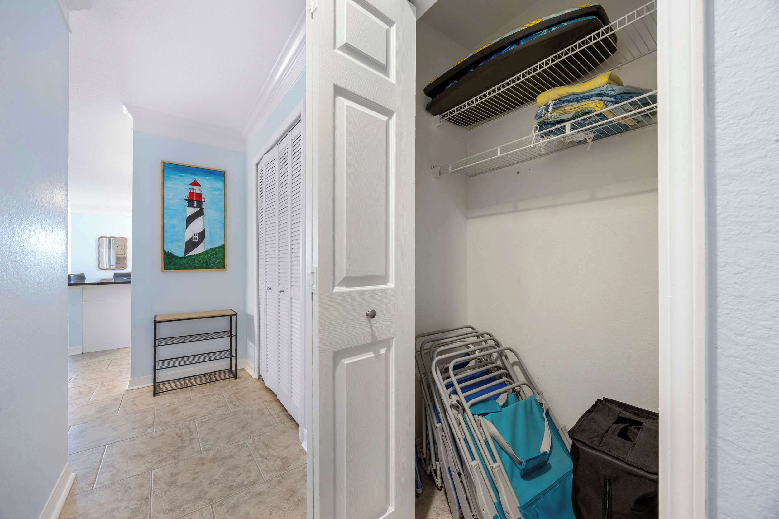Een gang met een deur naar een kamer, een schilderij van een vuurtoren, een radiator en een rek met opgevouwen kleding.