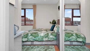 Ein Schlafzimmer mit Bett, Fenster, Pflanze und Blick auf Gebäude.