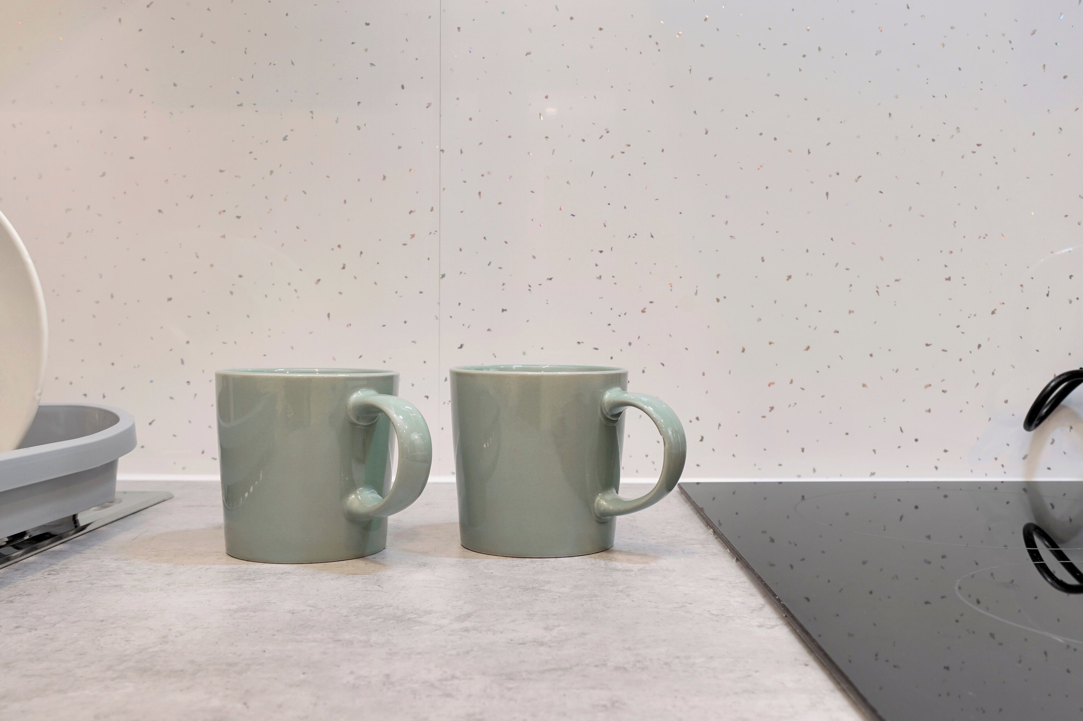Two light green ceramic mugs on a countertop with a speckled backsplash.