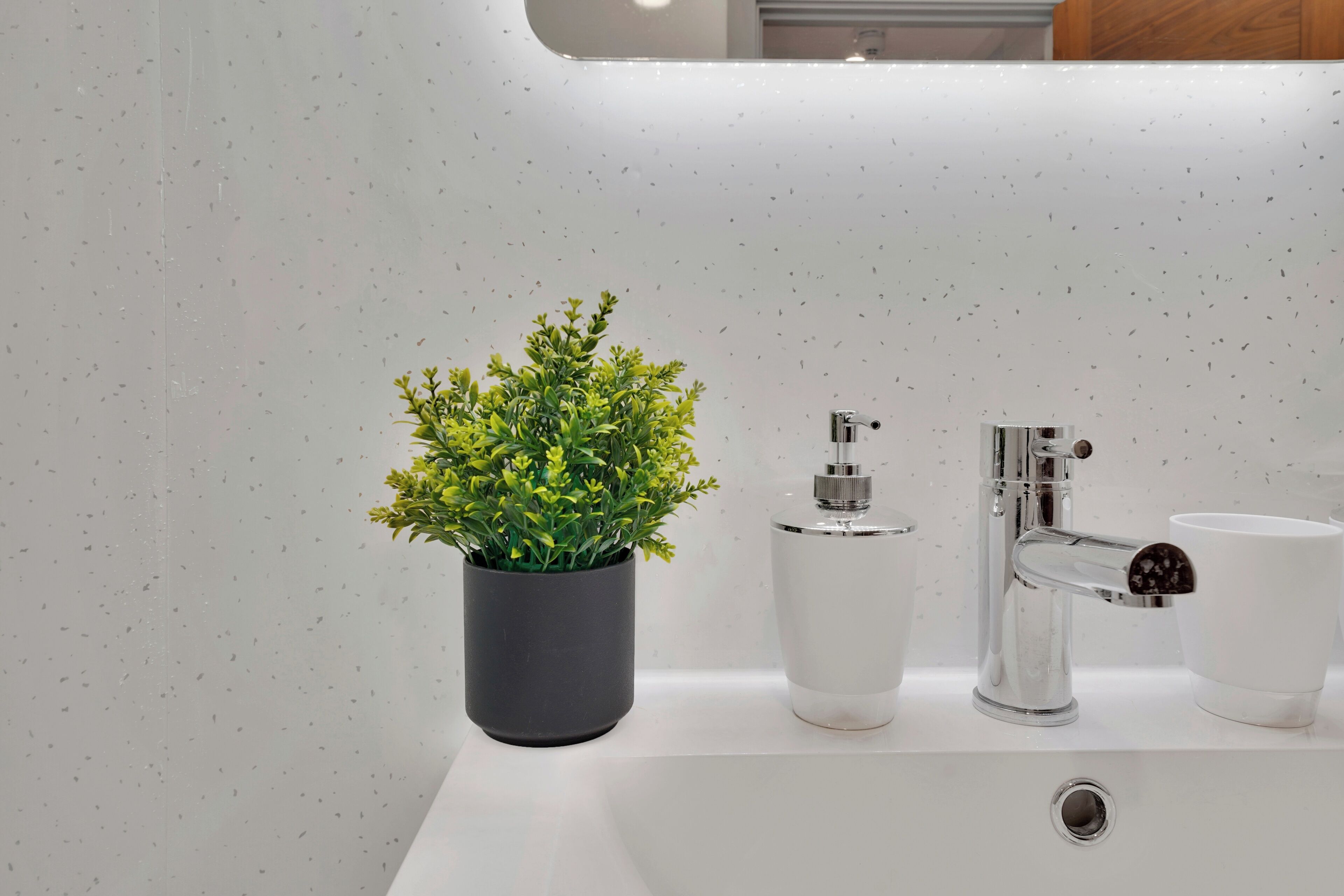 Une salle de bain moderne avec un lavabo blanc, une plante en pot, un distributeur de savon et une tasse.