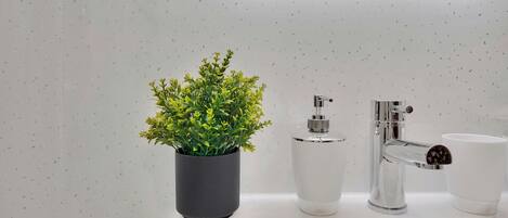 Une salle de bain moderne avec un lavabo blanc, une plante en pot, un distributeur de savon et une tasse.