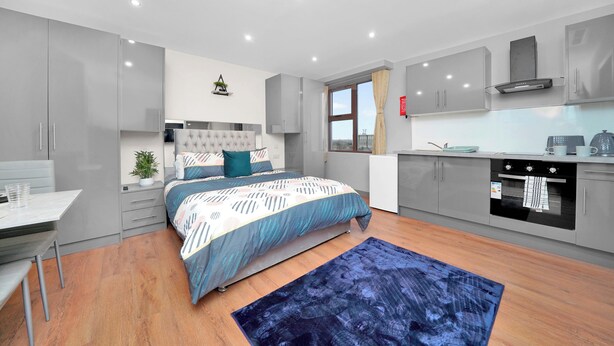 Une chambre moderne avec un lit, une table de chevet et un petit bureau. On y trouve une fenêtre avec des rideaux, une armoire intégrée et une kitchenette équipée d’une cuisinière et d’un four.