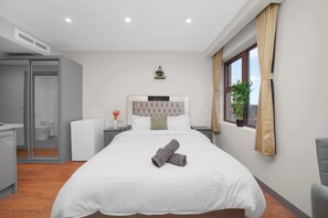 A modern hotel room with a large bed, a small desk, a glass shower enclosure, and a window with curtains.
