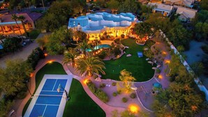 Una vista aerea di una zona residenziale con un campo da tennis, giardini curati e una grande casa con diverse sezioni.