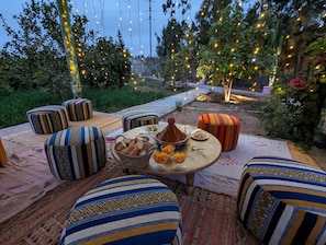 An outdoor dining area with striped cushions, a table with food, and string lights. - Dar Beynana (Sidi Bibi)