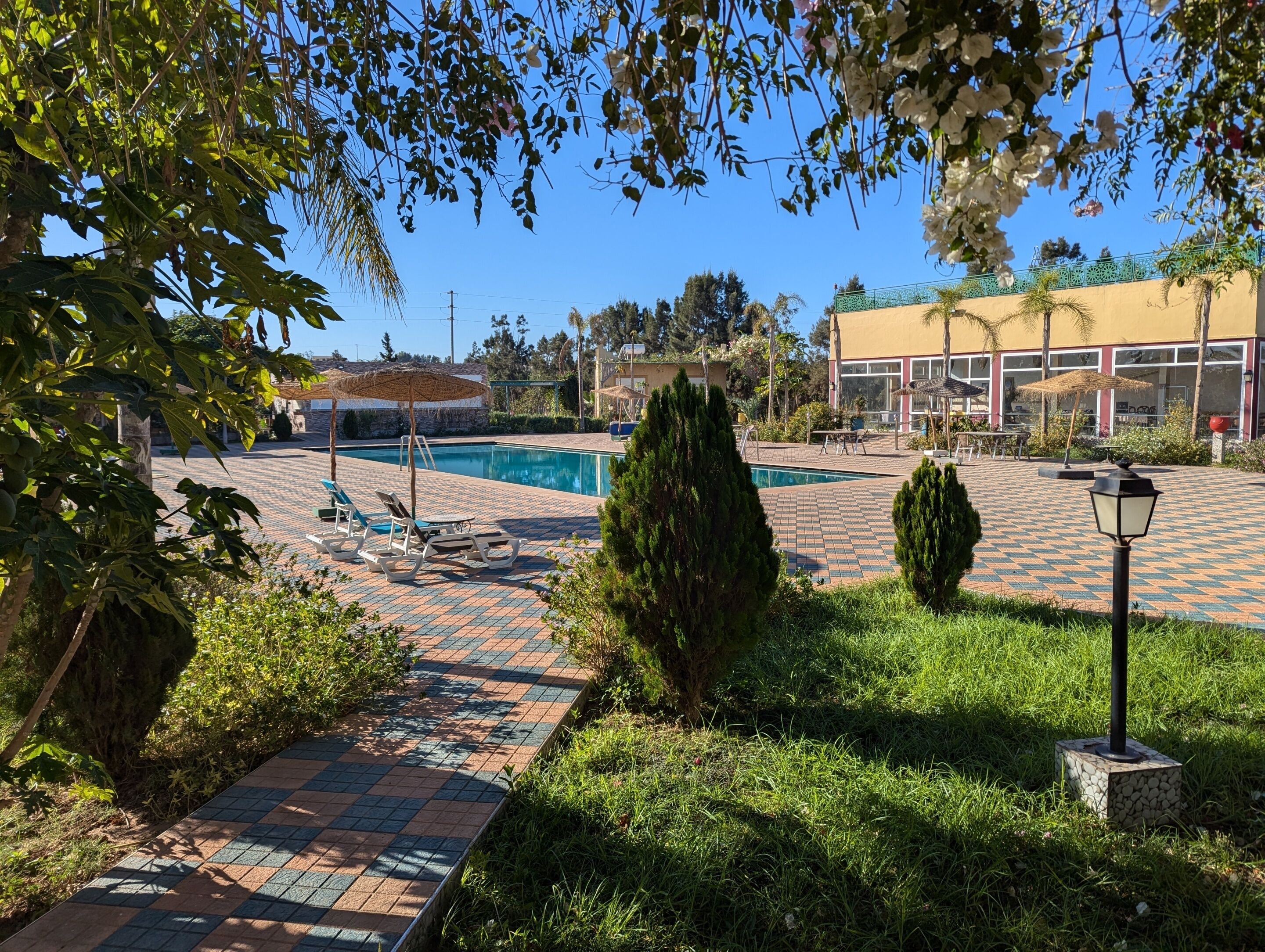 Un área de piscina con sillones reclinables, un camino empedrado y un edificio al fondo.