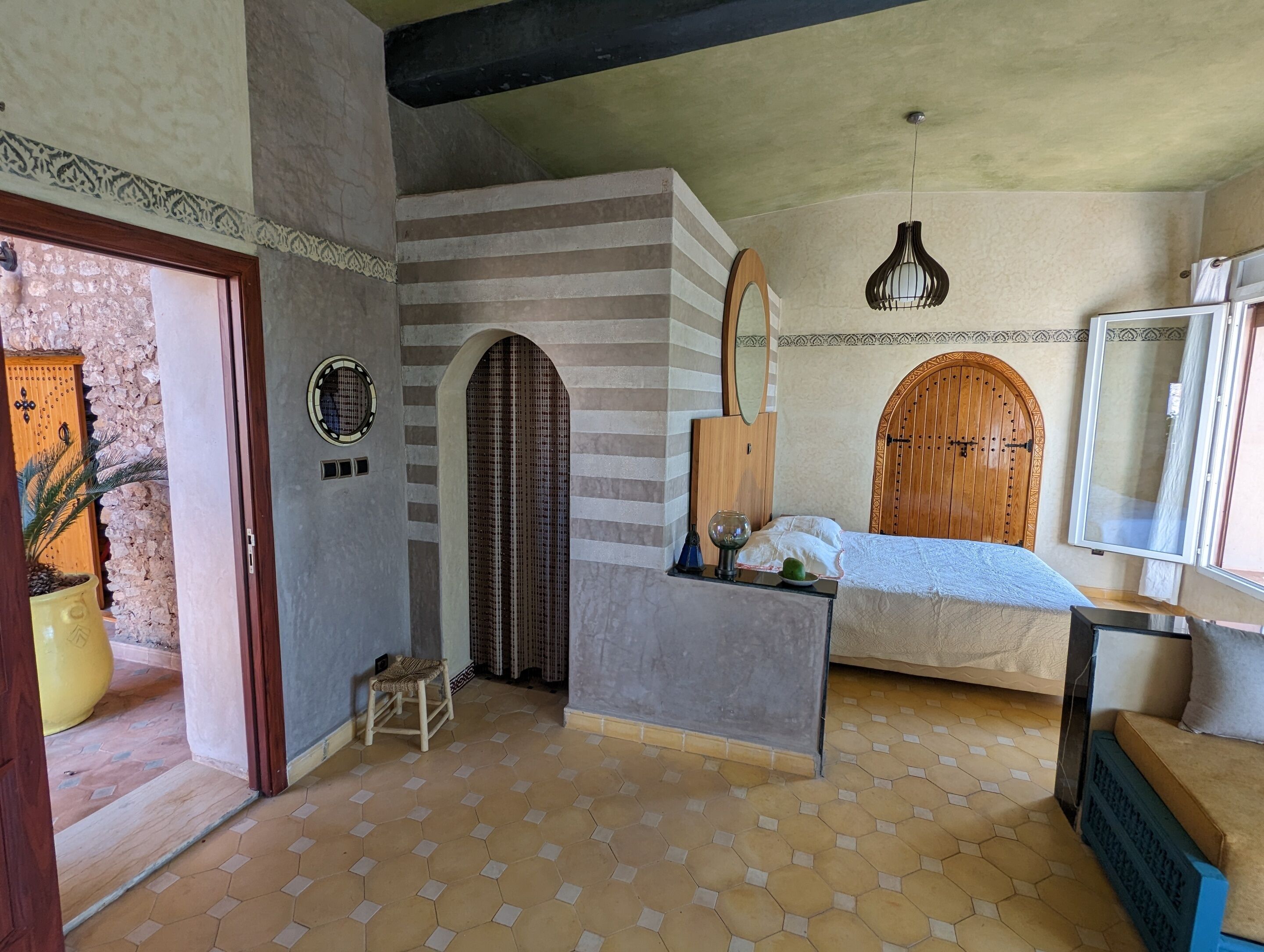 A room with a bed, a small stool, a wooden door, and a window.