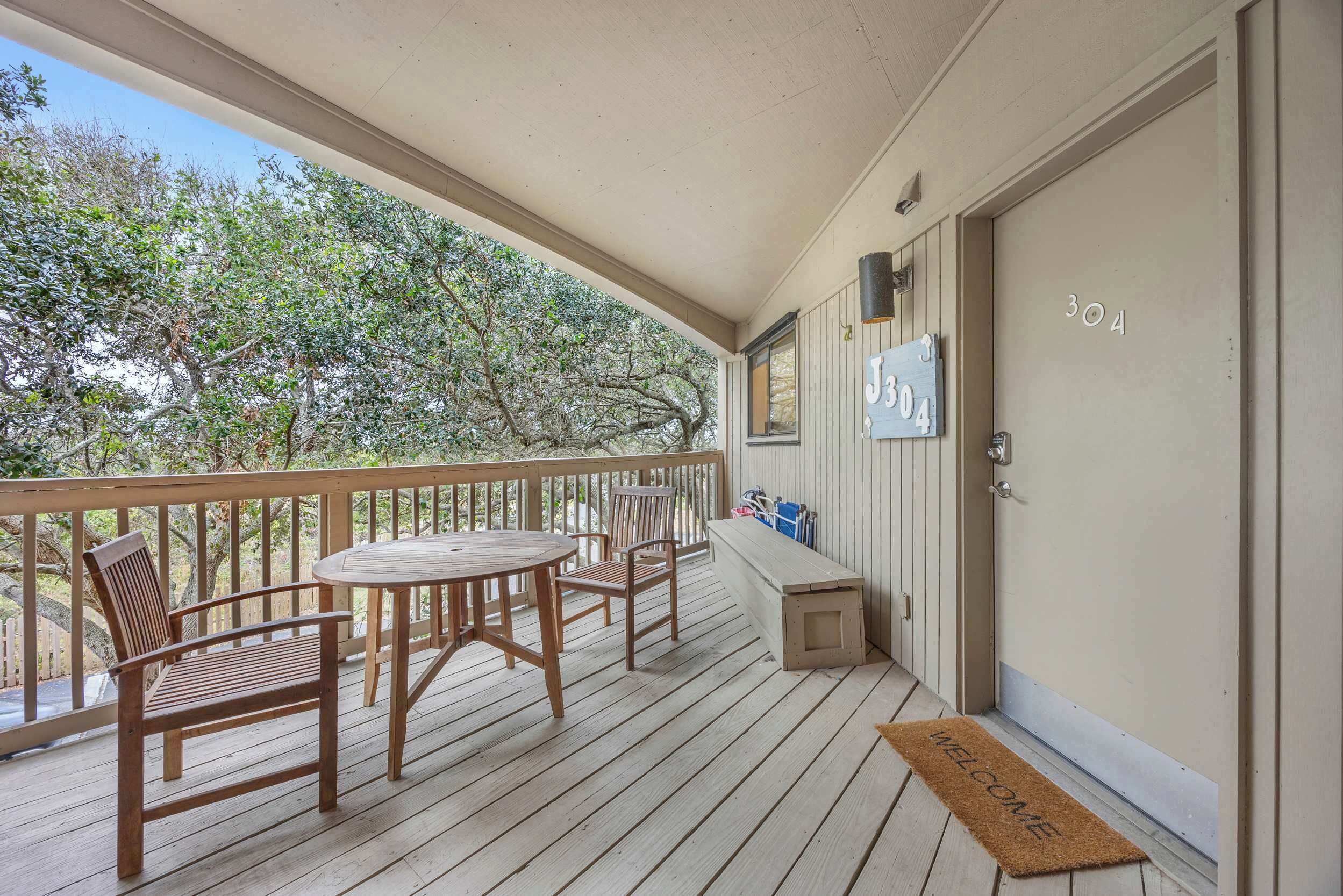 A wooden deck with a round table and chairs, a bench, and a welcome mat at the entrance.