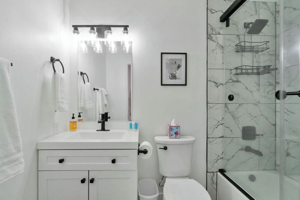 A modern bathroom with a white sink, mirror, and toilet. There is a framed picture on the wall and a shower with a glass door.
