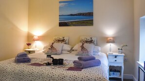 A neatly made bed with two pillows, a bedside table with a lamp, a small nightstand with a lamp, and a painting of a coastal scene on the wall. - 3 bedroom accommodation in Roch (Roch)