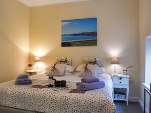 Cama bem arrumada com duas almofadas, mesa de cabeceira com abajur, mesinha de cabeceira com abajur e um quadro com uma paisagem costeira na parede.