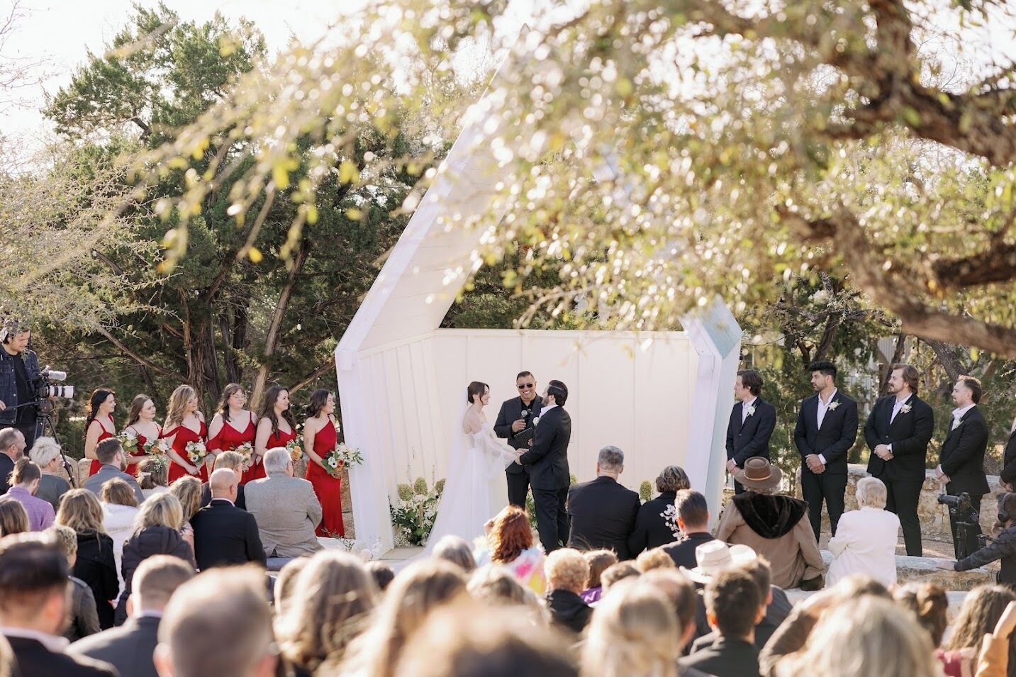 A wedding ceremony outdoors with a white arch and guests seated in rows.