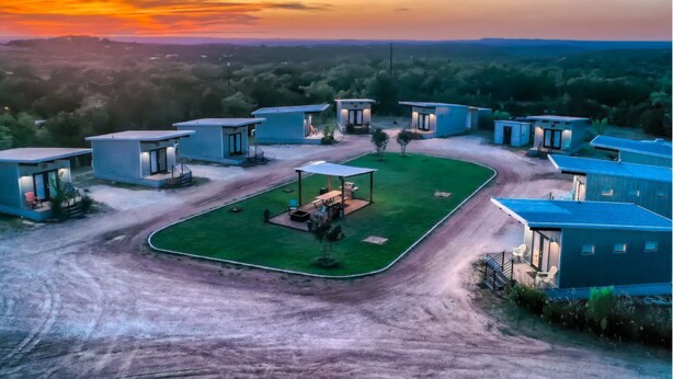 A modern resort with multiple blue-roofed cabins, a central open area with seating, and a dirt road leading to the cabins. - The Liney Moon (Dripping Springs)