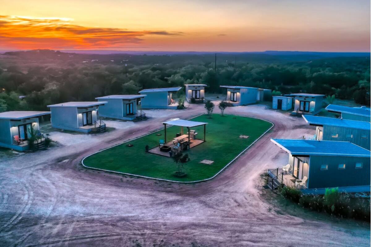 A modern resort with multiple blue-roofed cabins, a central open area with seating, and a dirt road leading to the cabins.