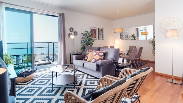 Un salon moderne avec un canapé gris, des fauteuils en osier, un tapis à motifs et un balcon offrant une vue dégagée.