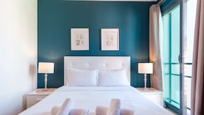 Un lit bien fait, avec du linge de lit blanc et des serviettes pliées, une tête de lit blanche, deux tables de chevet avec des lampes et deux reproductions botaniques encadrées accrochées au mur.