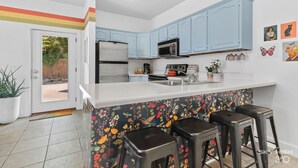 Een keuken met lichtblauwe kastjes, een wit aanrechtblad en een kleurrijke backsplash met bloemen. Er staan drie krukken bij het aanrecht.