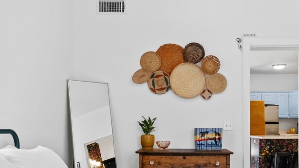 Een slaapkamer met een bed, een dressoir met een plant, een spiegel en een wand met gevlochten manden.