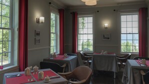 Een eetruimte met gedekte tafels, grote ramen met rode gordijnen en wandlampen.