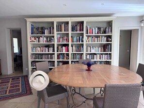 A room with a round wooden table, wicker chairs, a bookshelf filled with books, and a patterned rug. - Villa La Chaleureuse (Cabrières-d'Avignon)