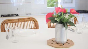 Een keuken met een wit aanrecht, een rieten stoel, een metalen kan en een vaas met bloemen.